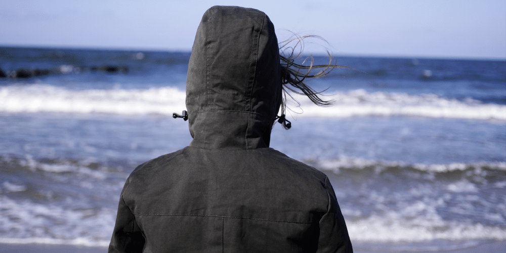 Person standing at the beach facing the water wearing a jacket with a hood.