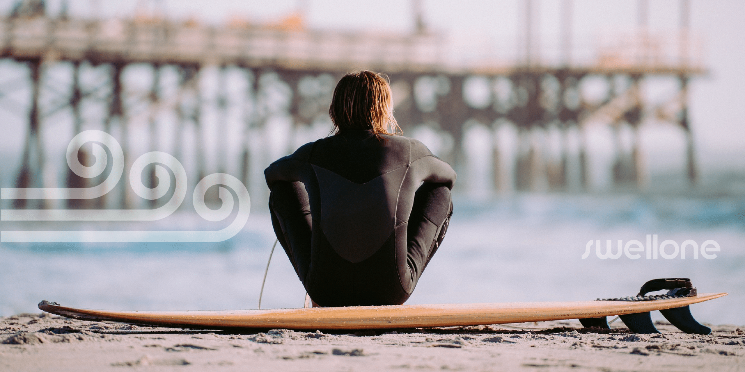 Surfer in wetsuit sitting at the water's edge with board